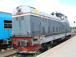 A navy-blueish locomotive with marine-ish topping depicted. Its registration reads 73 001-5. Buffer coupling and grabirons are visible in the sides. Driver cab is higher than long hood enabling two-way operation. There is a railcar painted in bright blue. Its window is open. Locomotive stays right by passenger platform and railcar in the track besides.