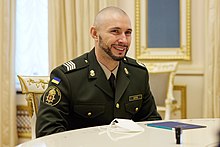 A man in a military uniform sits in an ornately-decorated room, smiling