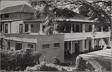 Black and white photograph of a three-storey building with wide verandas on the first and second storey. In front of the building is a cistern and it is flanked on the left by bushes and on the right by an overhanging tree.