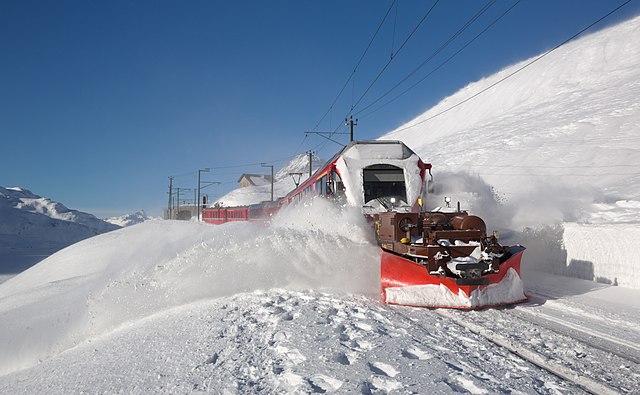 Электропоезд Rhaetian Railway ABe 8/12 Ретийской железной дороги с четырьмя прицепными вагонами регионального поезда R5 «Санкт-Мориц — Тирано» толкает перед собой снегоочиститель после выезда со станции Оспицио Бернина в районе перевала Бернина