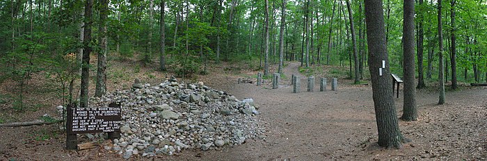 Panorama fotografico del "Memoriale di Thoreau" a Walden Pond.  A sinistra: pannello di legno con una citazione di Thoreau, tratta da Walden, poi un mucchio di pietre bianche depositate dai visitatori in sua memoria.  A destra e sullo sfondo: stele che delimitano il vecchio sito dove Thoreau costruì la sua capanna.  Tutt'intorno: alberi e tre sentieri che partono dal sito.