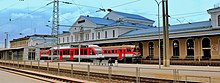 Seitliche Farbfotografie eines Bahnhofs mit einem Bahnsteig und Gleisen im Vordergrund. Das helle Bahnhofsgebäude hat Rundbogenfenster und die Aufschrift „Vilnius“. Vor dem Gebäude stehen zwei rote Züge und eine Person sitzt auf der Bank des Bahnsteigs, das vor den Zügen ist.