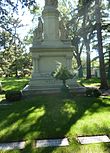 Большой каменный памятник на зеленом кладбище с двумя женщинами, смотрящими в разные стороны, и срезанной наверху фигурой. Из трех видимых могил Т. Б. Уокер находится в центре, а Гарриет Хьюлет Уокер - справа.