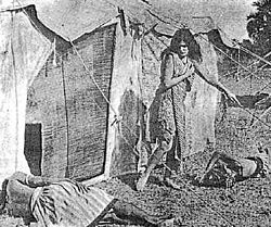Een jonge vrouw met lang donker haar loopt buiten een tent en kijkt neer op een van de twee mannen die op de grond slapen.  Ze draagt ​​alleen een sjaal en een knielange jurk, haar armen, onderbenen en voeten bloot.