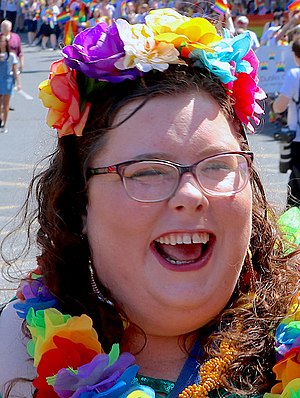 A close up photo of a white women wearing multicoloured floral headband, multicoloured floral garland, and rectangle glasses. She is laughing. She has long hair, and through the hair, you can see glimpse of her dangly earrings. The photo was taken on a sunny day. There are may people in the background walking, with rainbow coloured flag, and banner.