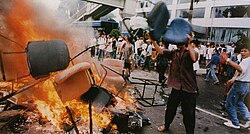 Um homem vestindo uma camisa abotoada, calças e chinelos joga uma cadeira de escritório em uma pilha de outras cadeiras em chamas no meio de uma rua da cidade.  Atrás dele, várias dezenas de pessoas se reúnem em frente a um prédio com janelas quebradas.