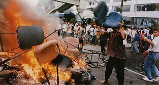 A man wearing a buttoned shirt, pants, and flip-flops throws an office chair into a burning pile of other chairs in the middle of a city street. Behind him, several dozen people gather in front of a building with broken windows.