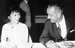 A middle-aged lady wearing a light-coloured dress and with short hair, fluffy at the front, sits at a dinner table smiling. To the right is a taller, older man in a dark suit, striped tie and light shirt who is turning his head to the left, talking to her. A man in a suit is visible, standing in the background.