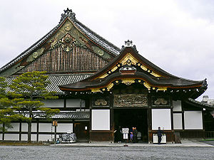 A small wooden structure with a hip-and-gable roof, white walls and metal ornaments on the gable is located in front of a large building with hip-and-gable roof, white walls and chrysanthemum motifs on the gable.
