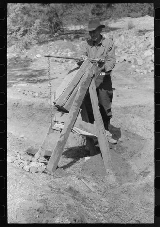 Prospector operating a drywasher, called a papago. The rocks and dirt are placed in the higher compartment. Below and to the back is a bellows made of canvas. This is pumped to blow through the screening and thus blow away loose dirt while the heavy gold remains. Pinos Altos, New Mexico, 1940. Papago dry washer.tiff