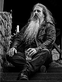 black-and-white picture of long-haired, long-bearded White male wearing a leather jacket, jeans, and boots, sitting on stone steps with a plastic cup of beer in his left hand