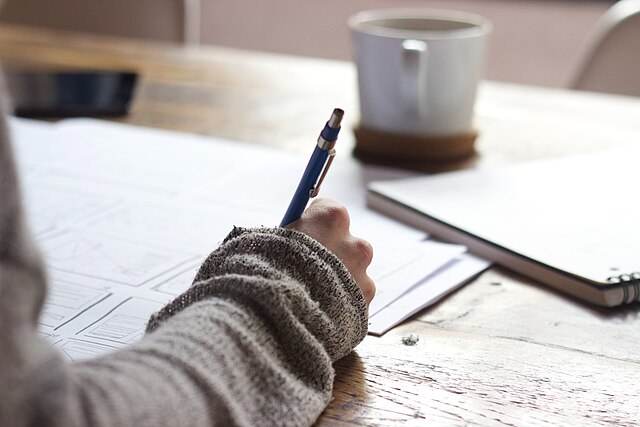  A person writing on a brown wooden table near a white ceramic mug