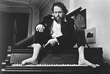 Schickele seated barefoot atop a piano, c. 1980s, pointing to his right foot which is deliberately playing the four notes that spell the name Bach: B-flat (which during Bach's time was simply referred to as 'B'), A (using his heel), C, and B (which during Bach's time was referred to as 'H').