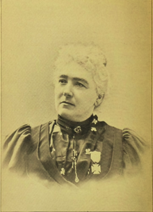 B&W portrait photo of a woman with her white hair in an up-do, wearing a high-collared dark blouse, with adornments, as well as a medal pinned onto it.