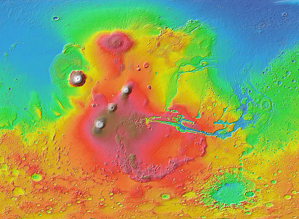 The Tharsis region (shown in shades of red and brown) dominates the western hemisphere of Mars as seen in this Mars Orbiter Laser Altimeter (MOLA) colorized relief map. Tall volcanoes appear white. The Tharsis Montes are the three aligned volcanoes left of center. Olympus Mons sits off to the northwest. The oval feature in the north is Alba Mons. The canyon system Valles Marineris stretches eastward from Tharsis; from its vicinity, outflow channels that once carried floodwaters extend north.