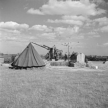 Black and white photo of a 3.7-inch anti-aircraft gun with ack-ack bursts in the sky above.