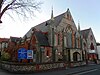 Front view of a wide three-part flint church with red brick dressings. The main (left) section is flanked by stone spires and has a large four-light lancet window with quatrefoils and a sexfoil, and two single-light lancets. In the middle is a narrow, low entrance section. To the right is a plain-walled section with three lancets and an arched window.