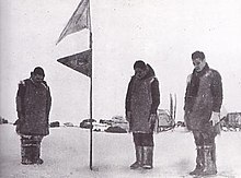 Photographie en noir et blanc de trois hommes saluant tête baissée un drapeau du Japon.