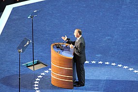 (A) The first three teleprompters: A monitor screen partly-embedded in the lectern's desk top displays the scrolling text of the speech in synchrony with two screens embedded in the podium floor. They are on either side of the speaker, reflected by the angled glass teleprompters above them. Click this image and the ones below to see the four-teleprompter system more clearly.