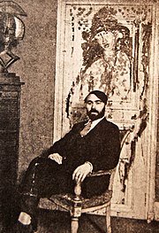 Photographie sépia d'un homme barbu assis dans un fauteuil, le dos à un tableau représentant un personnage.