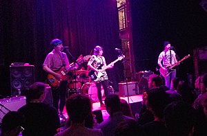 A dark, reddish-purplish image of a four-person band. To the left, a person with short blue hair and a button up shirt plays electric guitar. To the right, a person with medium brown hair, a dress, and leggings plays electric bass. Between and obscured by them, a drummer at a drum kit. To the far right, a person in a beanie, white T-shirt, and jeans plays electric guitar.