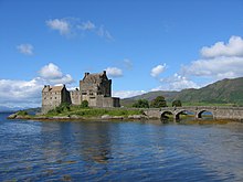 A stone castle in medieval Scottish style sits on a small island. An arched bridge leads off to the left.