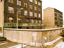 Top of the Wall with a smooth pipe, intended to make it more difficult to scale. The areas just outside the wall, including the sidewalk, are de jure East Berlin territory (1984). Exterior of East Berlin Neue Zeit newspaper, with Berlin Wall in foreground.jpg