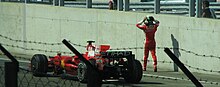 A figure in a red racing suit and boots, and wearing a helmet with a dark blue and light green design, walks away from a stationary red open-wheel racing car, which is parked next to the concrete wall separating the pit lane from the track. He is holding both his hands to his head. He has already removed his left racing glove.