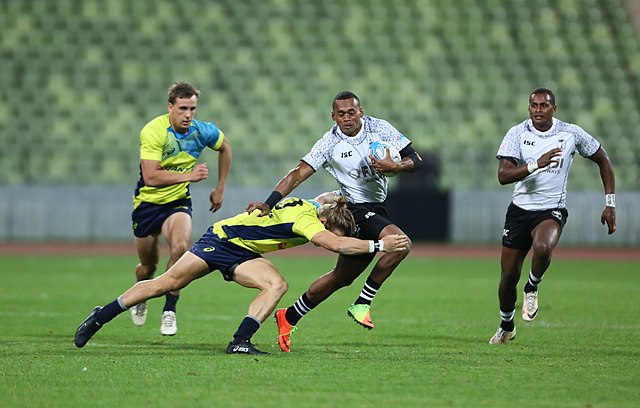 Tom Lucas (in yellow) attempts to tackle Samu Bale (in white), who is running with the ball towards the try line.