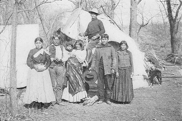 Photo of seven Apache men, women, and a child in front of lodges