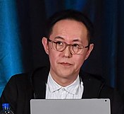 En middelaldrende japansk mann med briller, en hvit knapp og en svart frakk