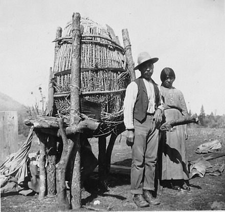 Mono people stand beside their Acorn cache in Fresno County, California, ca. 1920. Mono people used Acorns for their bread and families typically have 8 or 9 baskets of this size with acorns.[3]
