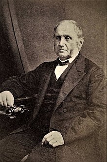 Sepia photo of middle-aged fair-skinned man sitting and half-turned to the viewer, wearing a three-piece mid-19th century business suit