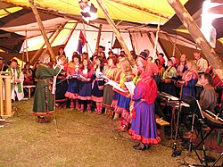 Vnitřek stanu, kde zpívá asi 30 lidí oblečených v sámských kostýmech. Vepředu je sbormistryně v zeleném oblečení.