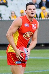 A male athlete with dark hair wearing a sleeveless jersey and shorts prepares to kick a football during a game of Australian rules football.