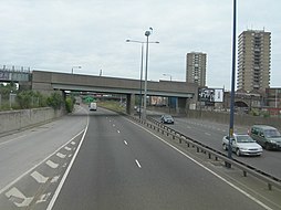 A dual carriageway crossed by a railway bridge in the foreground. Two high-rise blocks stand in the distance.