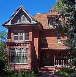 Ein reich verziertes Backsteinhaus mit einem links vorspringenden Giebelabschnitt und einem Walmdach im Hauptabschnitt dahinter. Die Türen, Fenster und die Veranda haben detaillierte Holzverkleidungen, die rot, gelb und grün gestrichen sind.