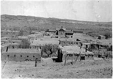 A black and white photograph of adobe buildings and a small church around a dirt plaza
