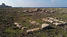 Sten spredt i et ret stort felt, nær ruinerne af et slot, ved havet.