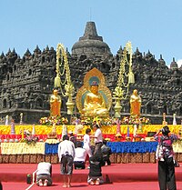 Asian devotees prostrating for images of the Buddha and two disciples. The images are positioned in front of a large stone monument (stupa).
