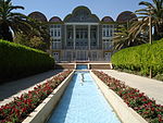 Water basin in a garden, flowers trees and a building with open portico.