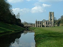 Uma fotografia de uma abadia em ruínas;  um rio passa na parte inferior esquerda da imagem, cercado por árvores escuras.  Uma construção de abadia em pedra em ruínas constitui o midground do lado direito da fotografia.