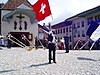 Au premier plan, un lanceur de drapeau en costume traditionnel; au second plan des joueurs de cor des Alpes en arc de cercle devant un calvaire; le cadre est une ville médiévale.
