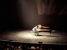Batiste reaching inside grand piano for special sonic effect during a lengthy improvised classical-jazz-avant garde opening piece in "Streams" performance in Red Bank, New Jersey. Jon Batiste reaching inside piano during improvised performance at Count Basie Theatre.jpg