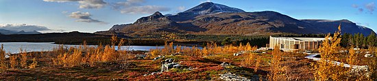 En bygning i en skog om høsten, nær en innsjø, med fjell i bakgrunnen.