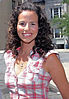 A sunny day, head-and-shoulders shot of a young lady with curly long dark hair, wearing a checked shirt, with a street in the background