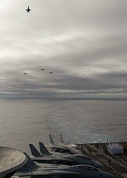 Photographed from the flight deck of an aircraft carrier: three airplanes are flying in formation while approaching. And a fourth plane is: climbing up. And away from the formation——to signify the 