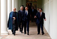 Quattro uomini, Obama al centro, parlano mentre camminano lungo una serie di colonne bianche.