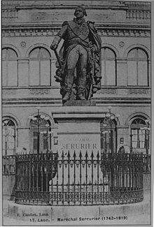 Fotografía antigua de una estatua de cuerpo entero de un mariscal de Napoleón en uniforme completo, sosteniendo su sable en su mano izquierda y su bastón de mariscal en su mano derecha.  Alrededor del pedestal, una rejilla, y al fondo, un edificio.