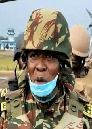A headshot of Valere Nka wearing a military camouflage helmet and camouflage jacket and body armour, as well as a bright blue surgical mask. there are two more soldiers in the background not wearing camouflage.